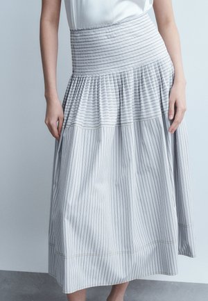 Γυναίκα με λευκή μίντι φούστα με λεπτές κάθετες μαύρες ρίγες και ζώνη στη μέση με σούρες, συνδυασμένη με λευκό αμάνικο τοπ.