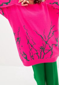 Rosa stickad tröja med gröna marmorliknande grafiska mönster, med en avslappnad passform och ribbade ärmslut, i kombination med gröna byxor.