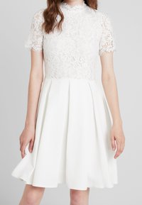 Femme portant une robe blanche avec un haut à manches courtes en dentelle et une jupe évasée, se tenant contre un fond gris clair uni.