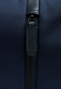 Dunkelblaues, strukturiertes Stofftaschen-Detail mit einem schwarzen Reißverschluss und einem Leder-Zipper und Akzent. Nahaufnahme des Materials und der Hardware.