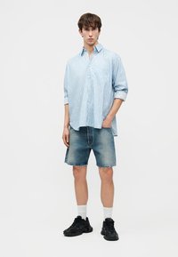 Jauns vīrietis, tērpies gaiši zilā svītrās pogājamā krekla, džinsa šortos, baltās zeķēs un melnos sporta apavos, stāvot pret vienkāršu baltu fonu.