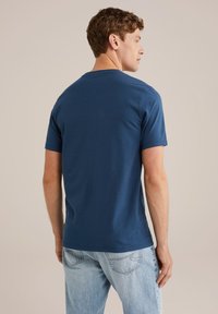 Tricou din bumbac de culoare bleumarin, cu mâneci scurte și gât rotund, purtat pe dos, asociat cu blugi din denim de culoare albastru deschis.