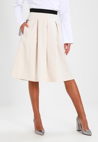 Jupe beige plissée avec une taille élastique noire, longueur genoux, dotée de poches latérales et d'une texture en coton lisse.