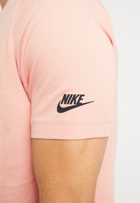 Camiseta de algodón de color rosa claro con un logo negro de Nike en la manga derecha. Diseño de manga corta y cuello redondo. Textura suave.