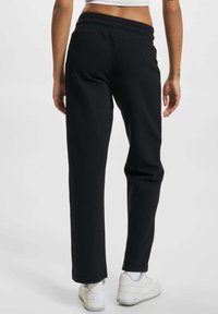 Czarne spodenki dresowe o luźnym kroju, z elastycznym paskiem w talii. W połączeniu z białymi trampkami, widok z tyłu.