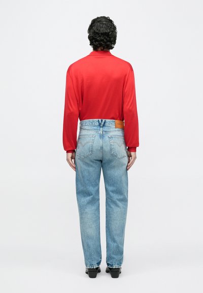 Mees lokkis tumeda juustega kannab punast pikkade varrukatega särki, helesiniseid teksapükse Versace sildiga ja musti kingi, seistes valge tausta ees.