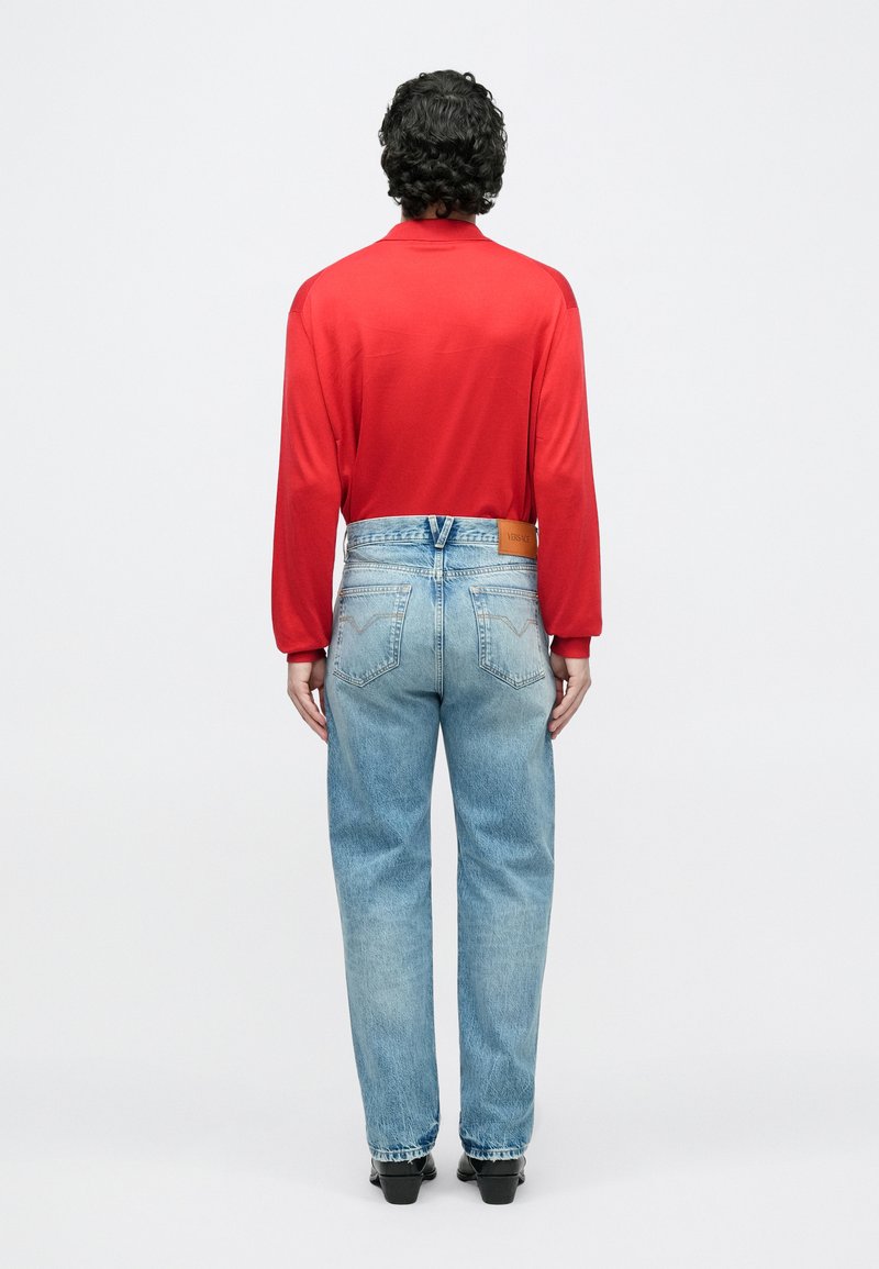 Man met krullend donker haar draagt rode lange mouwen shirt, lichtblauwe jeans met Versace-label en zwarte schoenen, staand tegen een witte achtergrond.