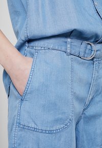 Ljusblå denimoverall med ett bälte med silverspänne, sidofickor och en avslappnad passform med subtila sömnadsdetaljer.