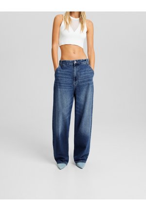 Femme blonde portant un crop top blanc sans manches et un jean bleu en denim ample, les mains dans les poches, avec des chaussures pointues bleu clair.