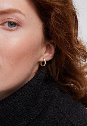 Złoty i srebrny kolczyk na uchu modelki. Kolczyk ma zaokrąglony kształt, gładką teksturę i błyszczące wykończenie.