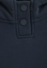 Mörkblått tyg med en jämn textur, med två mörka knappar märkta "POLO CLUB" nära kragen. Enkel design, inga mönster.