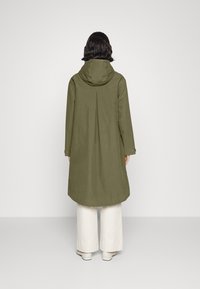Oliwkowy długi płaszcz z kapturem, z plisą z tyłu i przyciskami po bokach, noszony na beżowych szerokich spodniach. Gładka tekstura.