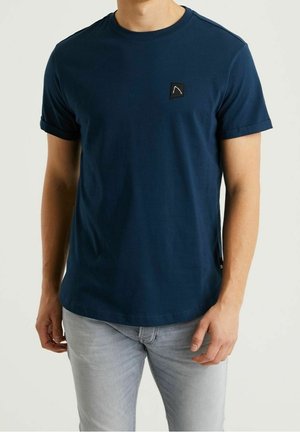 Mężczyzna ubrany w granatowy, krótki rękaw koszulki z małym czarnym kwadratowym logo na klatce piersiowej oraz jasnoszare dżinsy.