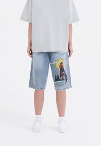 Bermudas de mezclilla con un lavado azul desgastado, que presentan un gráfico de un lobo contra una luna amarilla y el texto "NACIDO PARA SER SALVAJE" en una pierna.