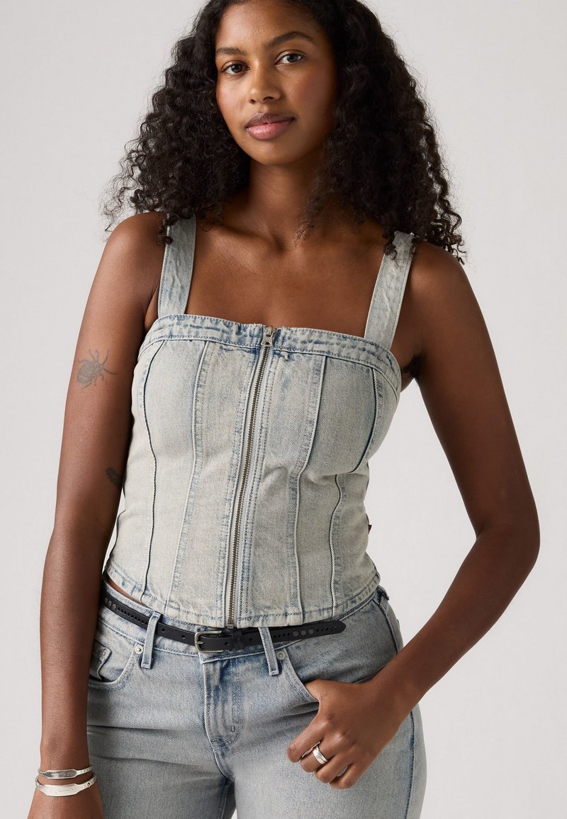 Donna con capelli ricci che indossa una giacca in denim chiaro con zip e jeans abbinati, con una cintura nera, bracciali d'argento e un anello.