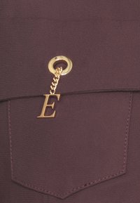Tessuto color marrone scuro con una piccola tasca, caratterizzato da un rivetto dorato e un accessorio a catena con la lettera "E" dorata. Texture morbida.