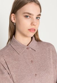 Camisa às riscas de cor rosa claro com gola e três botões. Textura suave com um padrão discreto. O modelo tem cabelo longo e brincos de prata.