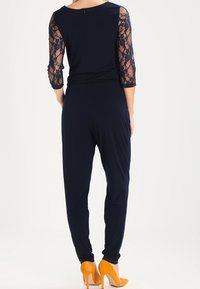 Marinblå overall med trekvartsärmar i spets, åtdragen midja och smala ben. Tillsammans med orange högklackade skor.