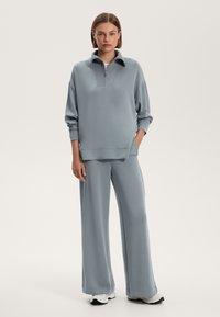 Svetlo modrá mikina s polovičným zipsom, voľný strih a dlhé rukávy, skombinovaná s rovnakými širokými nohavicami z jemného materiálu. Biely tenisky.