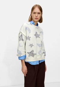Felpa color crema chiaro con motivi di stelle e lune grigi. Tessuto morbido con una vestibilità rilassata, indossata sopra una camicia azzurra con colletto.