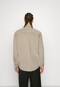 Lysebeige ribbet skjorte med lange ærmer, knapmanchetter og en buet kant. Har en plissédetalje bagpå.