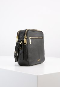 Schwarze, strukturierte Leder-Crossbody-Tasche von Fossil mit goldenen Reißverschlüssen und verstellbarem Riemen, auf einer weißen Fläche präsentiert.