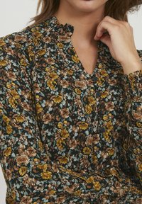 Florale Bluse mit dunklem Hintergrund und senfgelben, braunen und blauen Blumen. Rüschenausschnitt und Knopfdetails mit gerafften Ärmeln.