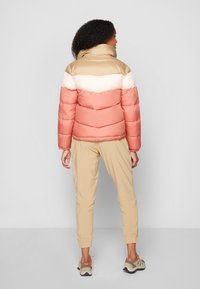 Puffjacka i persika, krämfärgad och beige, med hög krage, quiltad textur och elastiska mudd. Kombinerad med beige joggers och sneakers.
