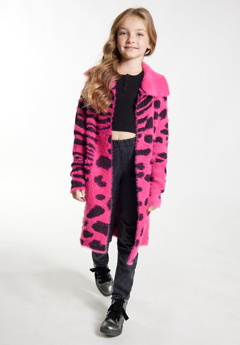 Roze i crni lažni krzneni kaput s uzorkom leopard, širokim ovratnikom, dugim rukavima i bočnim džepovima, nosi se preko crne majice i hlača.