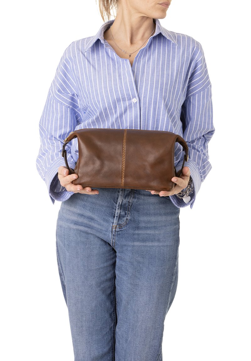 Borsa in pelle marrone di forma rettangolare, con una cucitura centrale e manici piccoli, tenuta contro una camicia a righe blu chiaro.