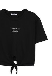 T-shirt nera in cotone con maniche corte, scollo rotondo e dettaglio anteriore annodato. Presenta un testo bianco: "CIÒ CHE NON È MIO NON È UN MIO PROBLEMA."