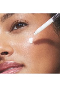 Une crème blanche est appliquée sur un visage lisse et hâlé. La peau a une texture brillante, et un applicateur transparent est tenu au-dessus.
