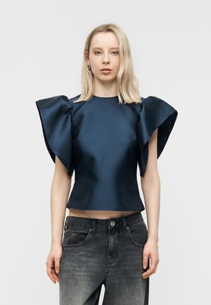 Sieviete, kas valkā tumši zilu augšu ar lielām volānu piedurknēm un izbalējušas melnas augsta vidukļa džinsas, stāvot pret vienkāršu pelēku fonu.