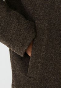 Detail hnedého plsteného saka s rukou vo vedľajšej vrecku, s textúrovaným povrchom a viditeľným šitím v blízkosti dolného okraja rukáva.