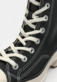 Juodos aukštos sportinės avalynės su baltomis raišteliais, metaliniais akyčių užsegimais ir kontrastingu grietinėlės spalvos guminiu padu. Yra matomos siuvimo linijos ir tekstilinis viršus.