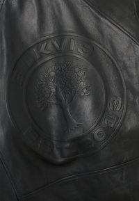 Czarna skórzana powierzchnia z tłoczonym okrągłym logo przedstawiającym szczegółowe drzewo oraz tekst „BLKVIS EST 2013.”