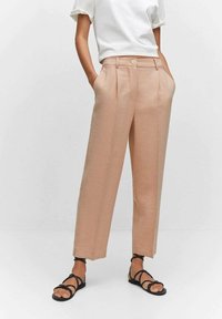 Pantalon beige sur mesure à taille haute avec des plis devant. Porté avec un haut blanc à manches courtes et des sandales noires à lanières.