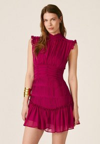 Mujer joven con cabello largo y castaño que lleva un vestido plisado sin mangas de color fucsia y una pulsera de brazalete dorada en su muñeca izquierda.