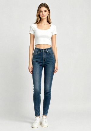 Giovane donna che indossa un top bianco corto, jeans skinny blu a vita alta e sneaker bianche, in piedi davanti a uno sfondo semplice.