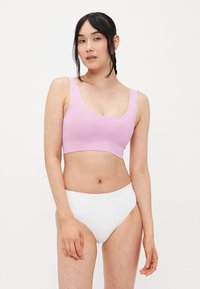 Žena s dugom tamnom kosom u pletenici nosi svijetlopurple crop top bez rukava i bijele gaće s visokim strukom, stojeći protiv jednostavne pozadine.