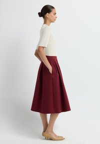 Jupe bordeaux mi-longue avec des plis, associée à un haut crème ajusté. La tenue comprend des poches latérales et des chaussures à talons nus.