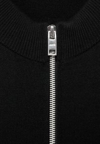 Acercamiento a un zipper de metal plateado en el cuello de una prenda de punto negro con canalé, zipper parcialmente cerrado.