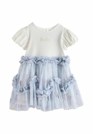 Ragazza sorridente con i capelli raccolti in uno chignon, che indossa una maglia bianca a maniche a sbuffo, una gonna di tulle azzurro chiaro con volant e scarpe bianche con grandi fiocchi, in piedi all'interno.