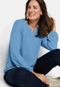 Maglietta a maniche lunghe blu chiaro con scollatura a V e texture a costine, abbinata a pantaloni scuri e sneakers chiare su una figura seduta.