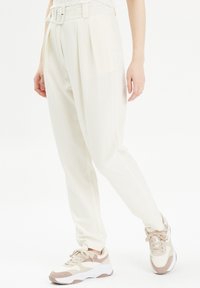 Pantalon blanc sur mesure à coupe large, avec détail de ceinture et tissu texturé, associé à des baskets beiges et blanches.