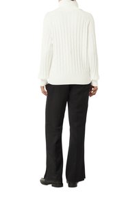 Weißer Cable-Knit-Pullover mit hohem Kragen, gerippten Manschetten und Saum; kombiniert mit schwarzen weit geschnittenen Hosen und schwarzem Schuhwerk.