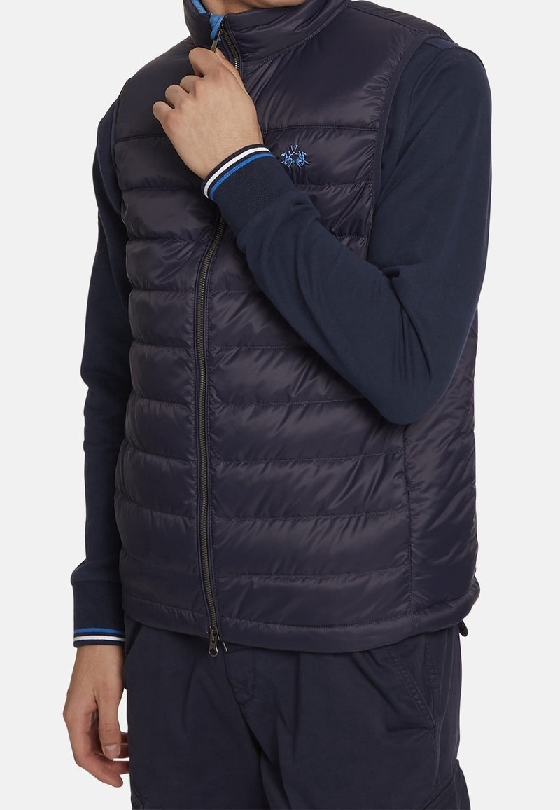 Navy puffer vest with horizontal quilted texture, full front zipper, high collar, and blue striped accents on the inner collar.