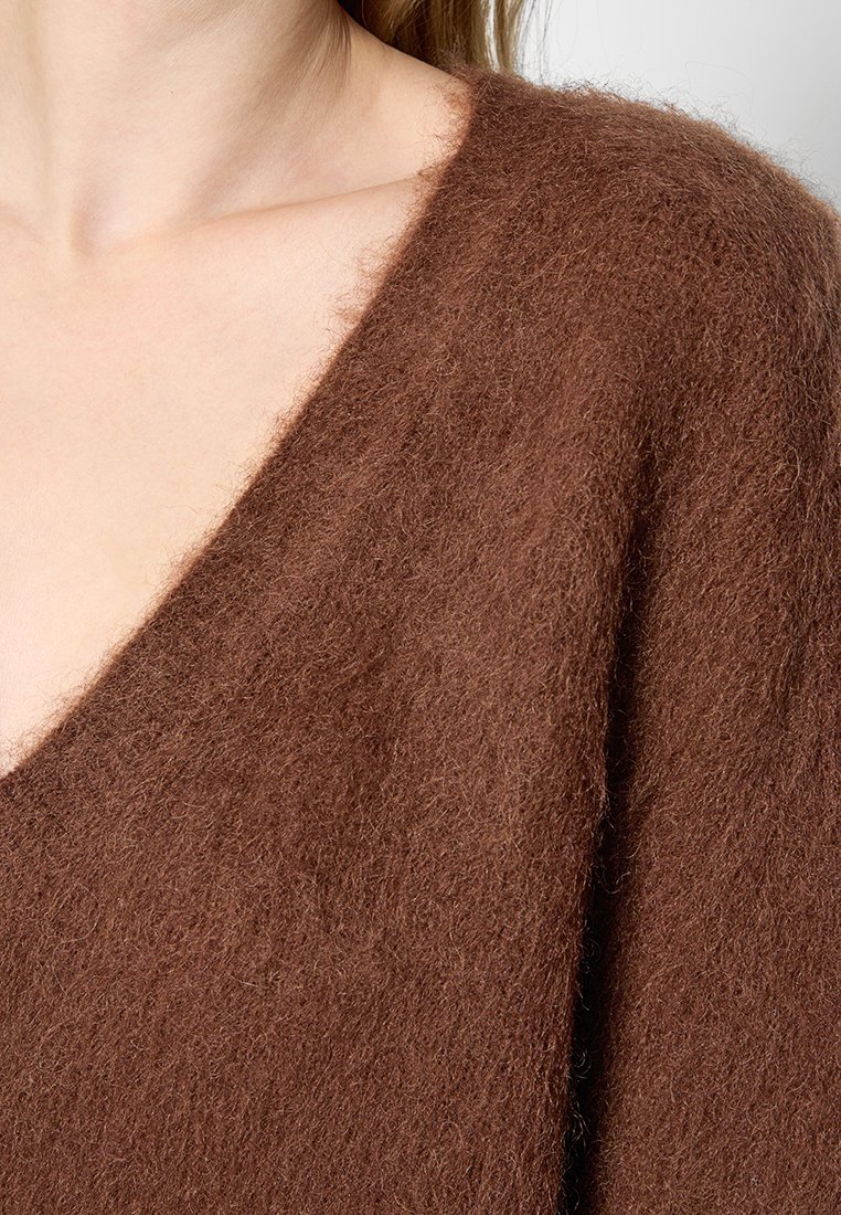 Femme portant un doux pull en laine brun avec un décolleté en V profond, laissant apparaître une partie du cou et du haut de la poitrine.