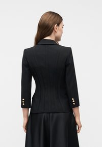Γυναίκα που φαίνεται από πίσω φορώντας ένα κομψό μαύρο σακάκι με χρυσά κουμπιά στα μανίκια και μία ταιριαστή μαύρη φούστα, με φόντο ένα απλό σκηνικό.