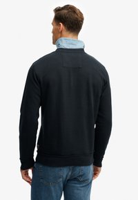 Sort sweatshirt med en struktureret overflade, der viser en blå skjorte med krave, der titter frem ved halsudskæringen. Synlige syninger og en detalje med et baglap.